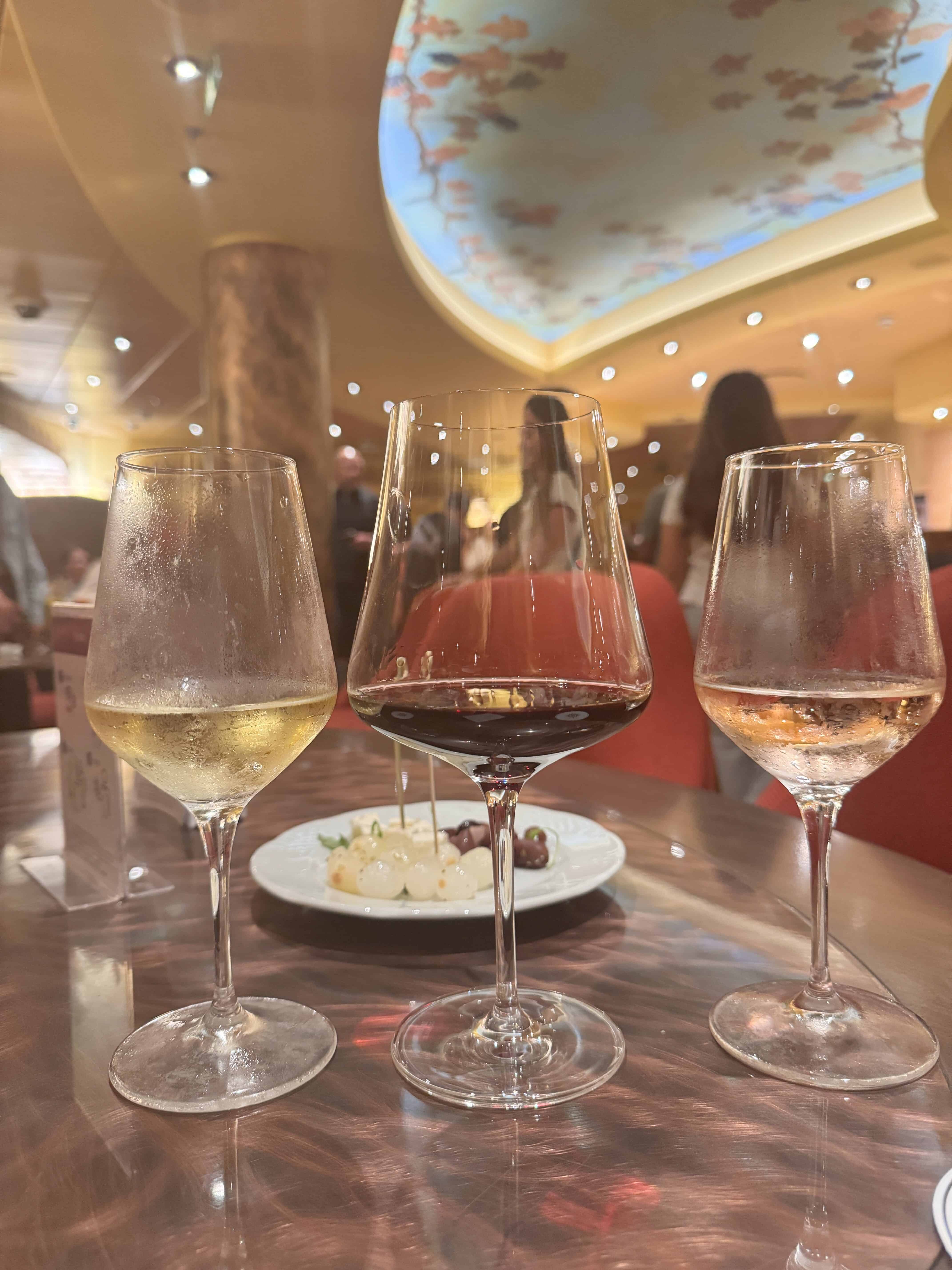 Three wine glasses—white, red, and rosé—on a table with a plate of food in a warmly lit restaurant, capturing the elegant dining experience you can expect aboard MSC Fantasia; perfect for your 2025 cruise review.