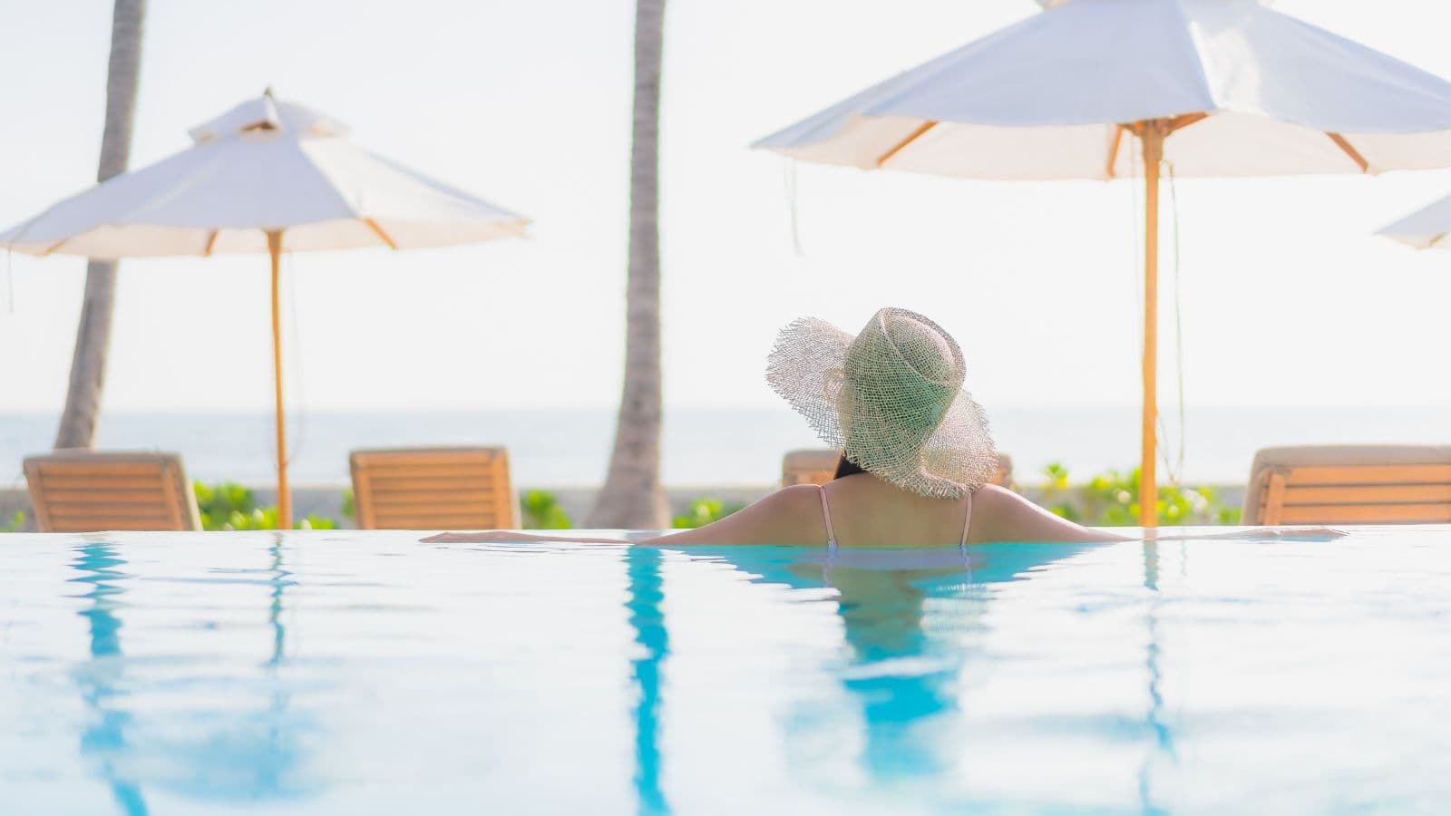 A person wearing a wide-brimmed hat relaxes in a swimming pool, facing away from the camera. Wooden lounge chairs and white umbrellas hint at why travelers love all-inclusive vacations by the beach or ocean.