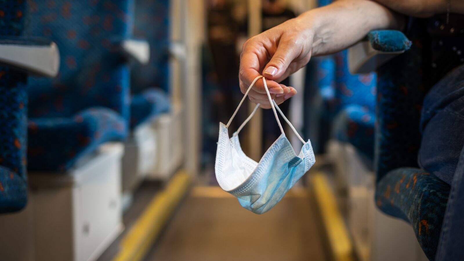 A person holds a disposable face mask by its ear loops while standing in the aisle during a quiet train ride with blue upholstered seats—a great moment to remember essential train travel tips.