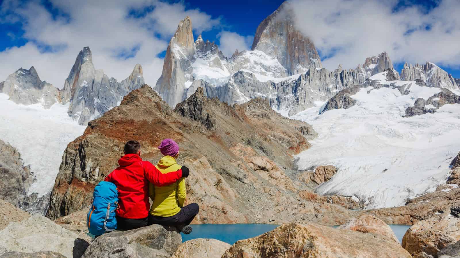 Two hikers sit on rocks, embracing, and admire snowy mountains and a blue sky in the background.