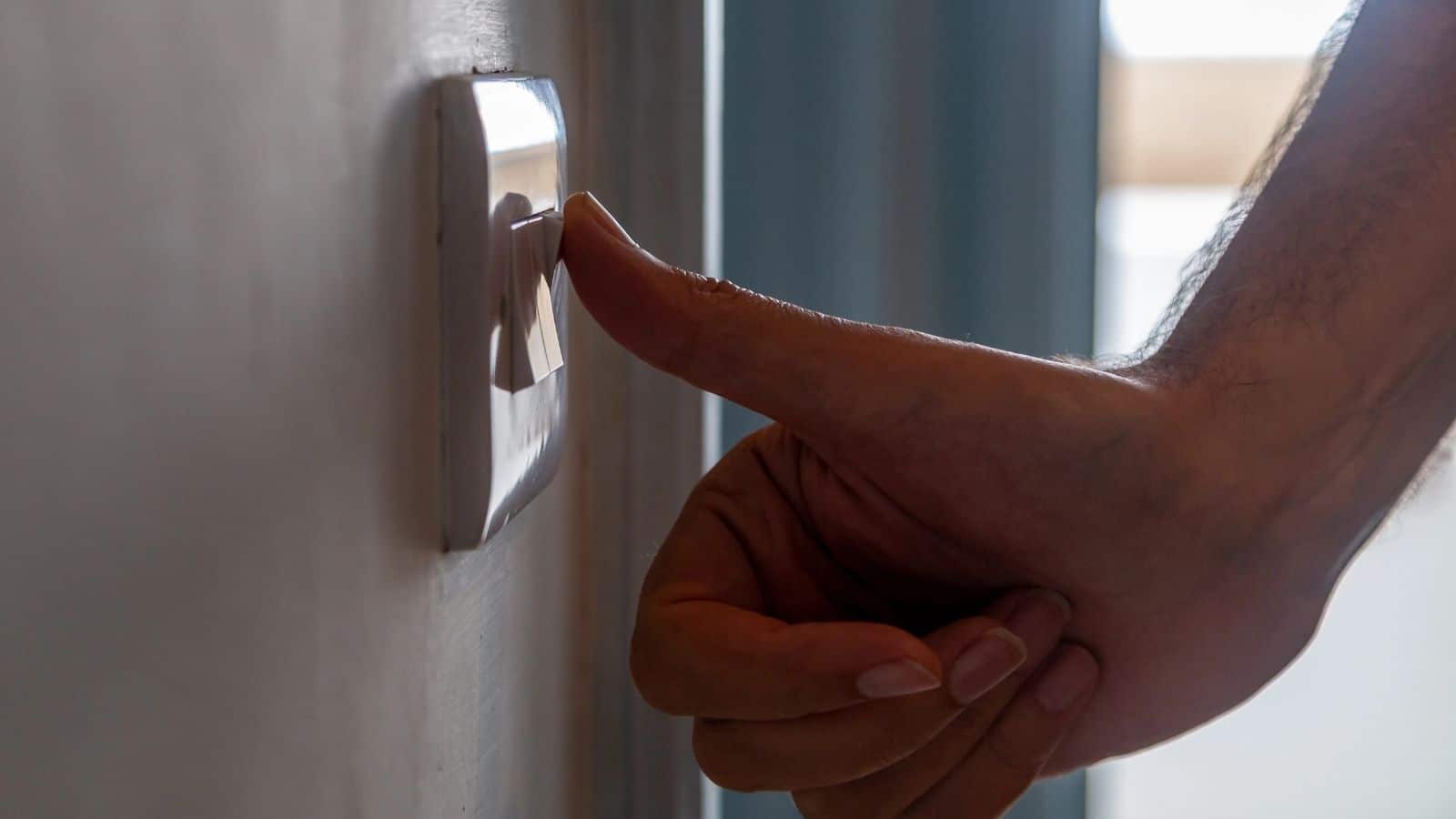 A person’s hand is pressing a light switch on a wall—an action that often causes travel confusion for first-time travelers unfamiliar with European habits. The scene is indoors, with the background softly out of focus.