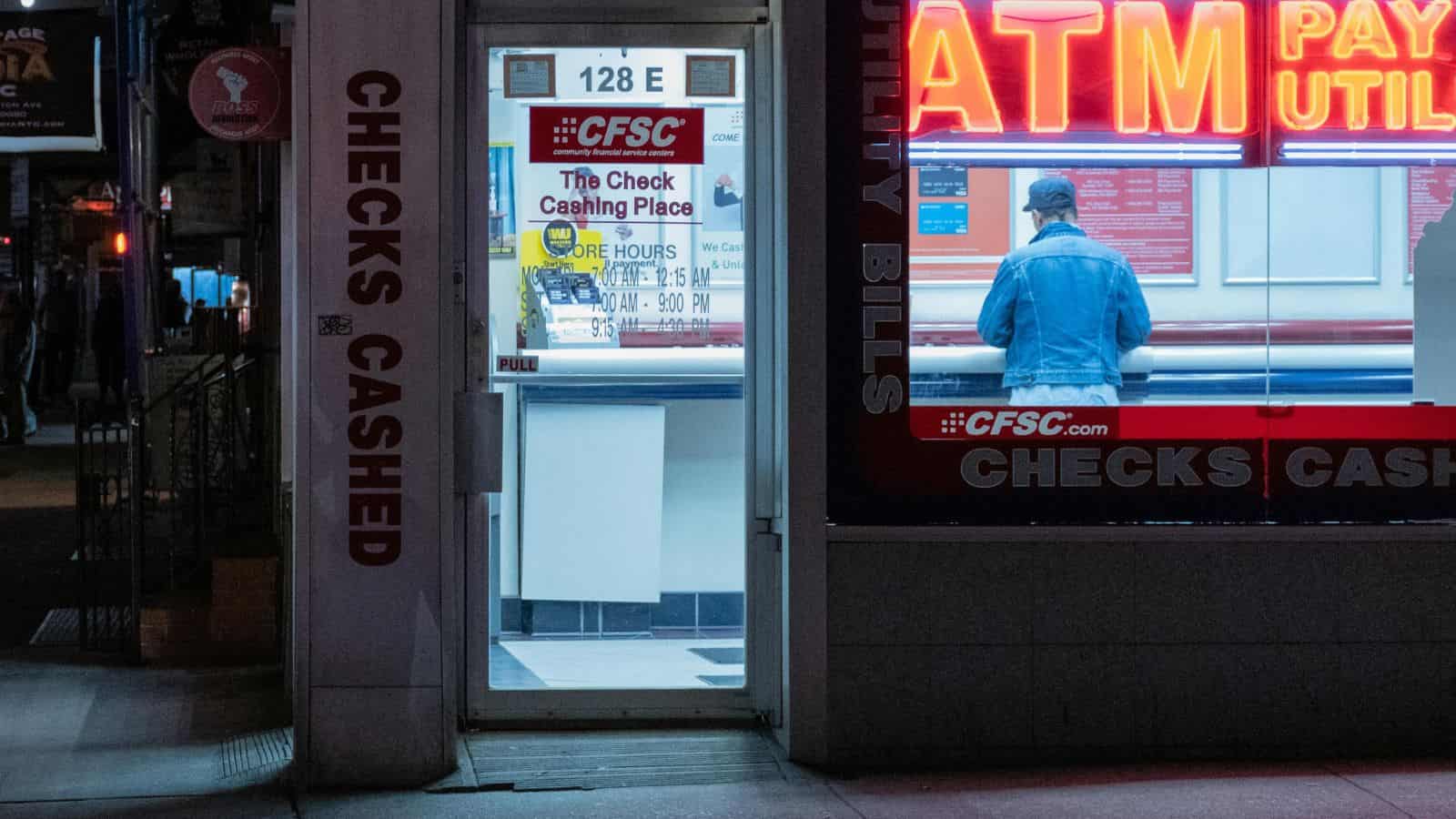 A person uses an ATM at night inside a brightly lit check cashing store.