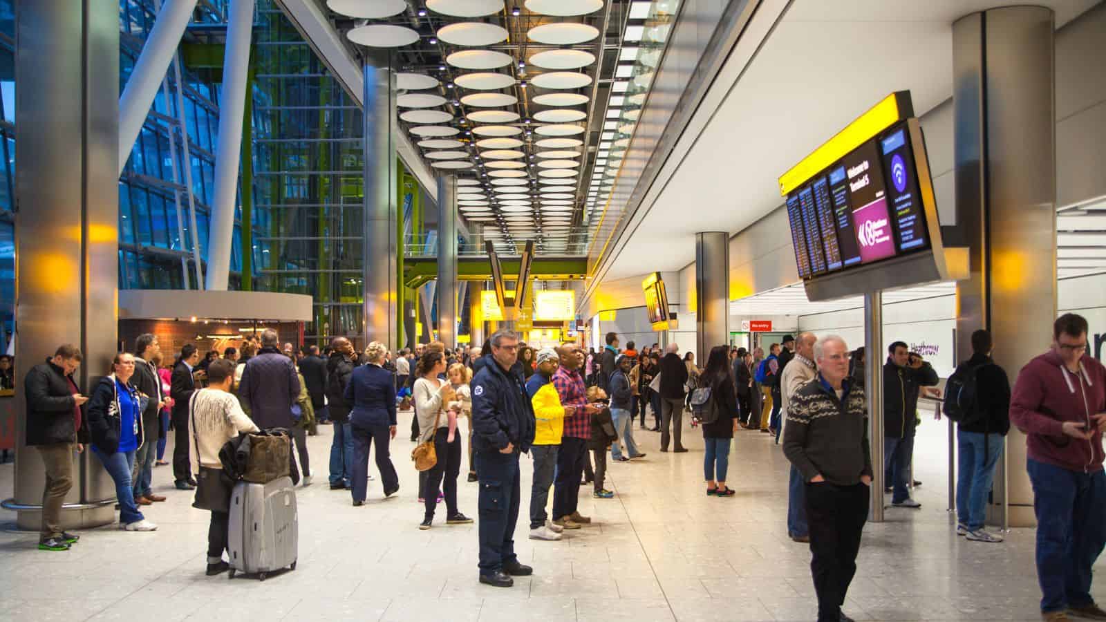 A busy airport terminal with passengers standing, some with luggage. Information screens and bright yellow signs hang from the ceiling. Large windows allow natural light into the spacious, modern interior.