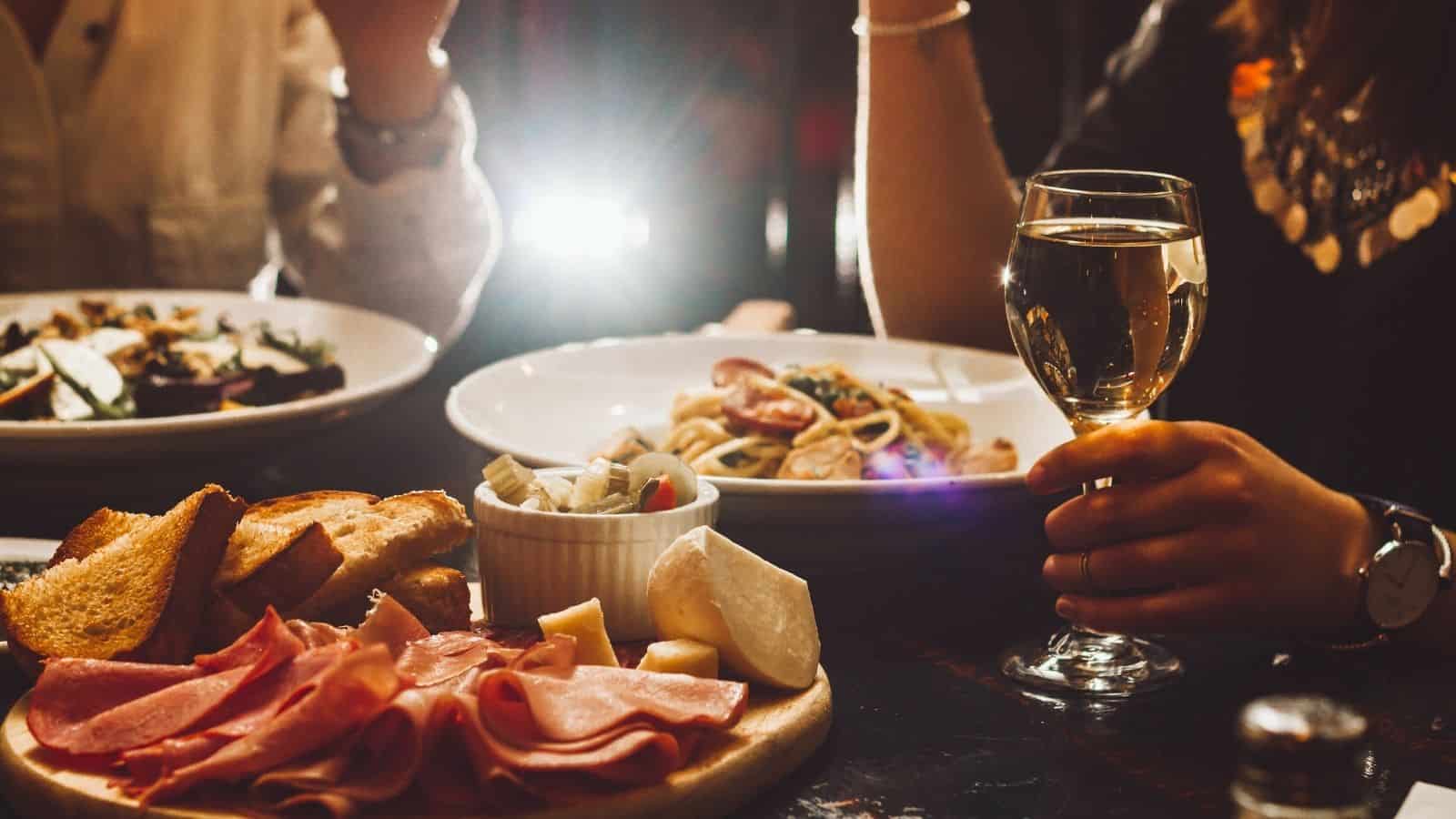 A person holds a glass of white wine at a table set with assorted cheeses, cold cuts, bread, and pasta dishes—capturing classic European habits that first-time travelers often seek to experience. Another guest is partially visible across the table.