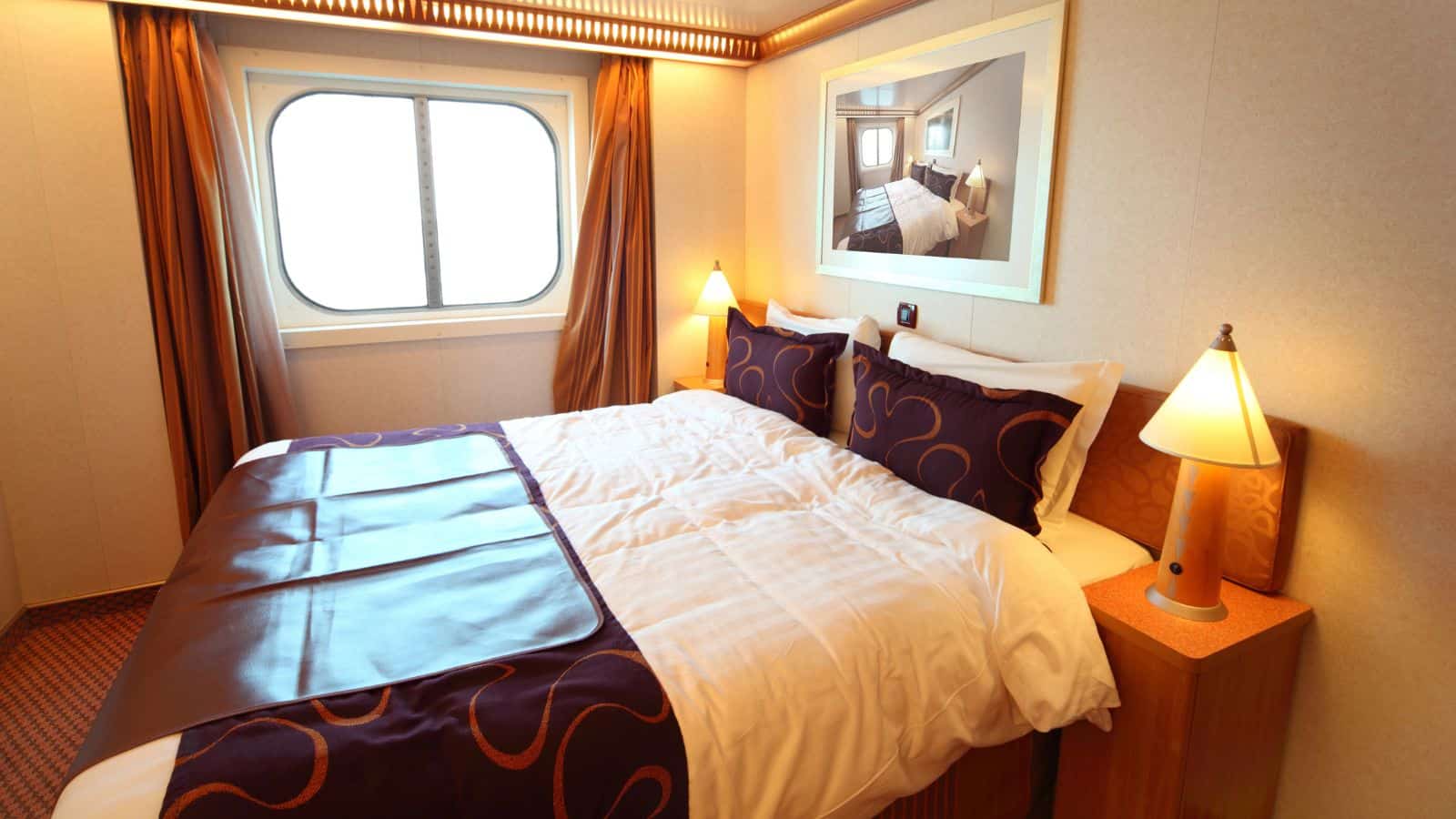 A neatly made bed with white sheets and a brown and purple runner, two bedside tables with lamps, a large window with curtains, and a framed picture above the bed in a compact, well-lit room.