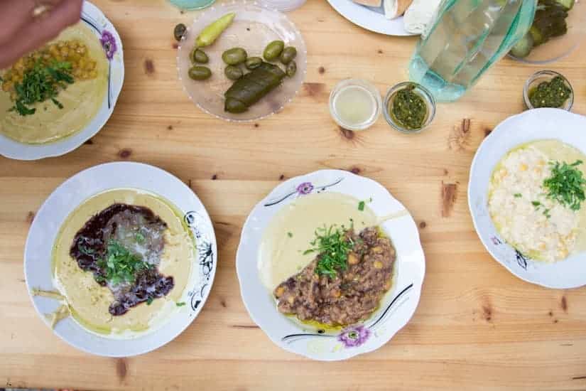 Wooden table with plates of hummus, beans, pickles, sauces, and a glass jug of water.
