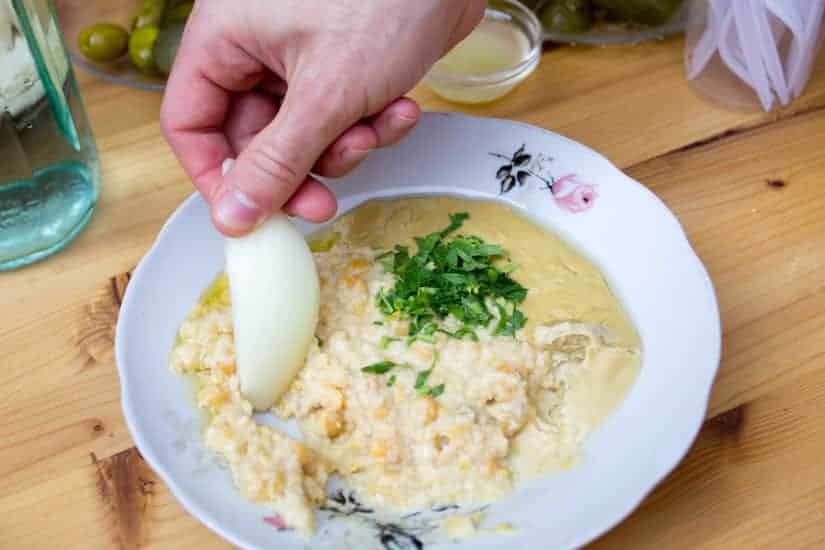 A hand dips an onion slice into hummus topped with chopped parsley on a white plate.