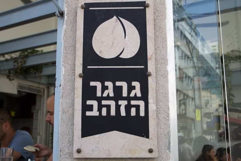 A black and white sign with Hebrew text and a fruit logo, mounted on an outdoor wall.