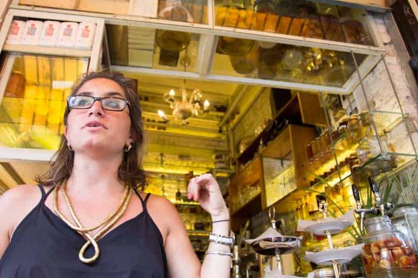 Woman in glasses stands inside a shop with shelves of jars and goods behind her.