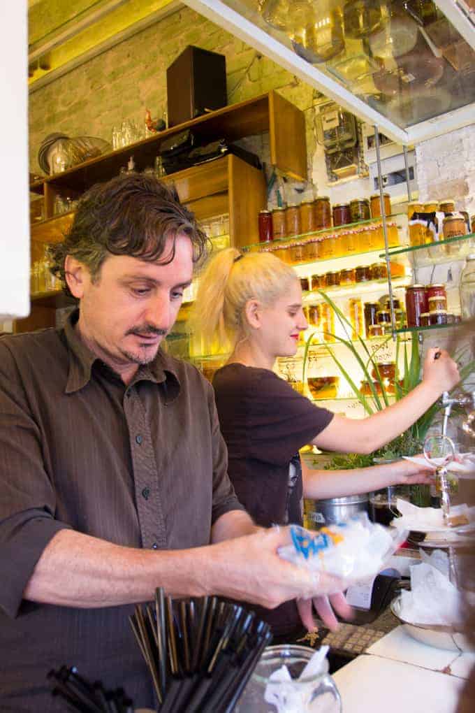 Two people work behind a busy café counter with jars on shelves and various kitchen tools around them.