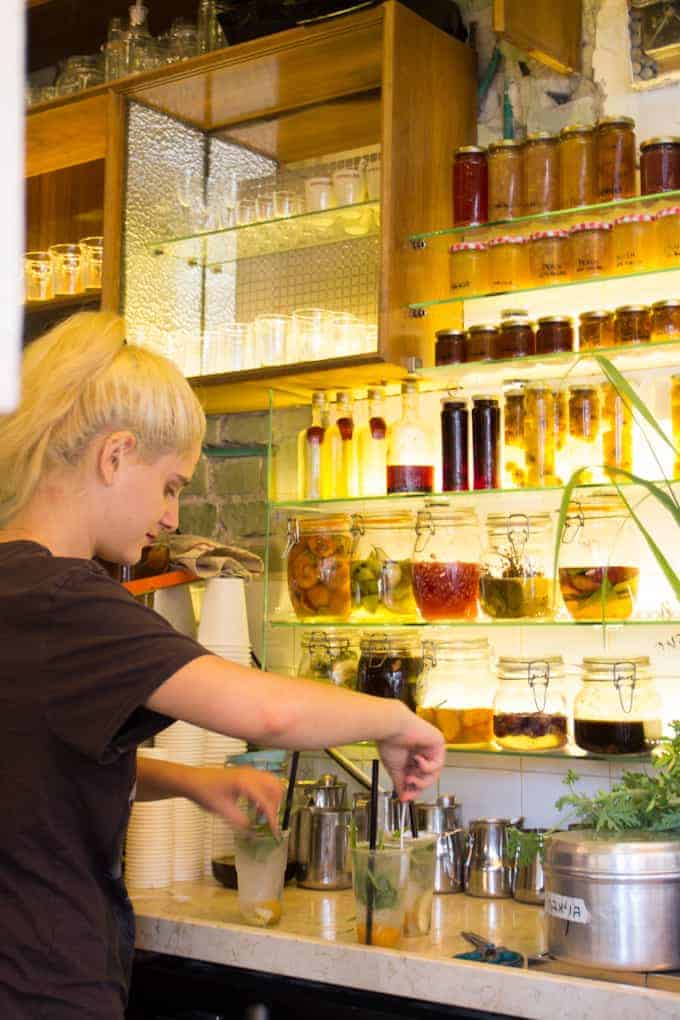 A person prepares drinks at a bar with shelves of jars, bottles, and glasses in the background.