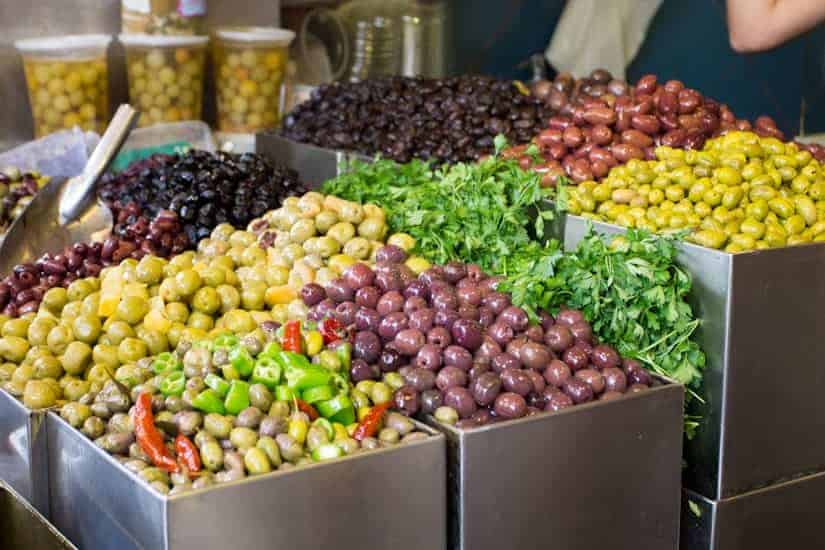 A market stall displays assorted olives in metal bins, with herbs and peppers mixed in.