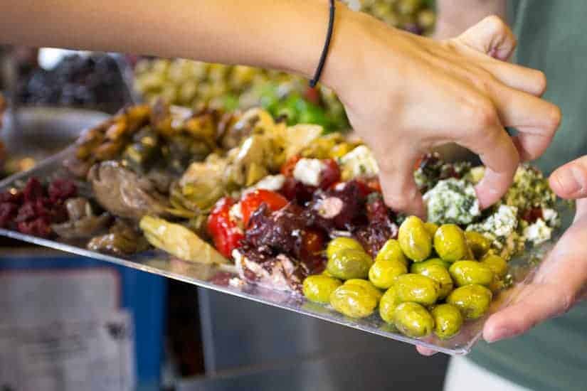 A hand reaches for assorted olives and antipasti on a tray at a market or food stand.