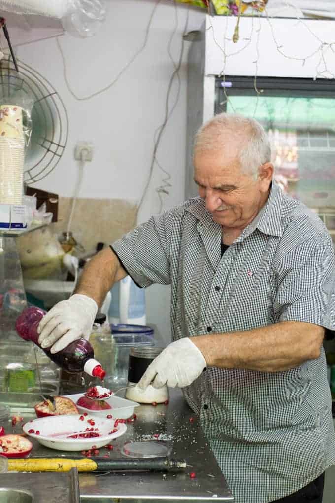 Older man in gloves squeezing pomegranate juice into a cup at a messy kitchen counter, smiling.