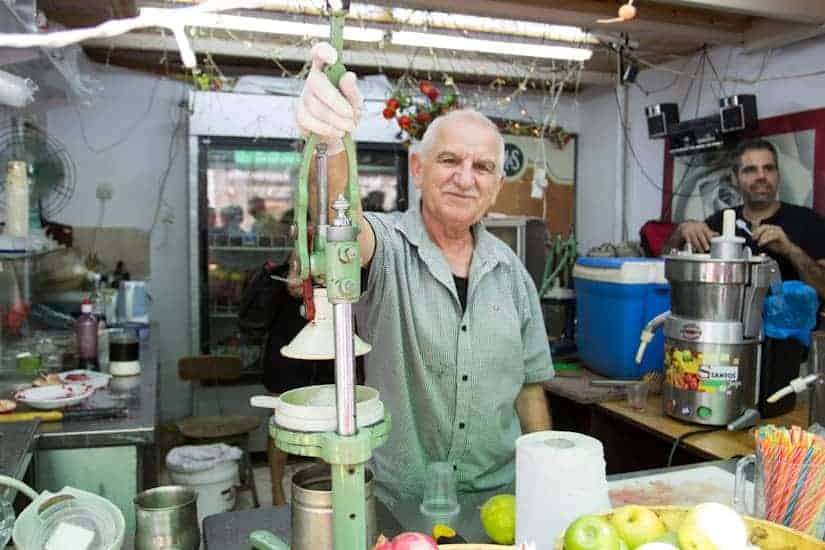 An older man smiles while making juice at a fruit stand, surrounded by fresh fruit and juicing equipment.
