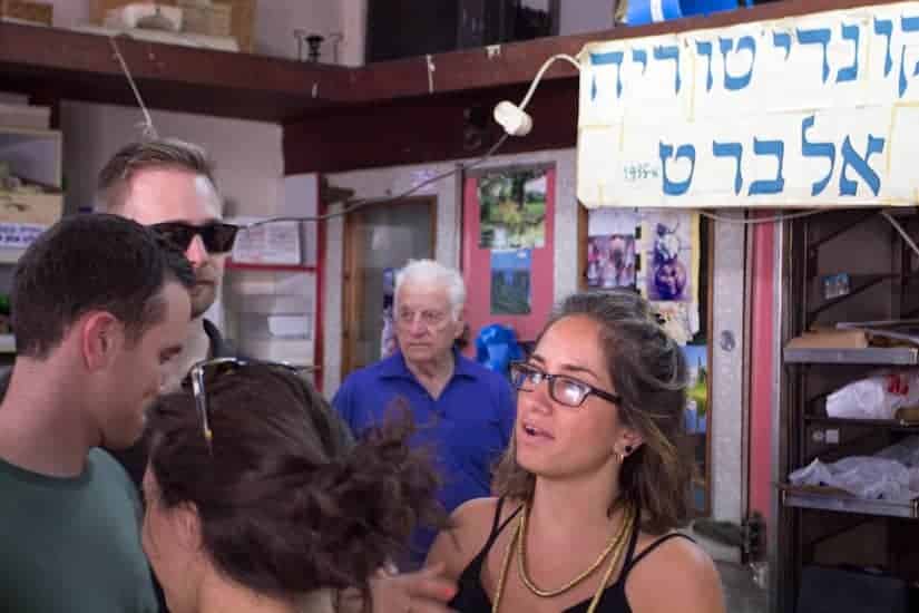 A group of people talk inside a shop; a man in blue stands in the background near a sign with Hebrew text.