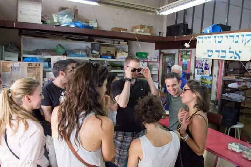 A group of people gathers and chats inside a shop with shelves and supplies in the background.