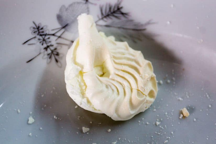 A partially broken white meringue cookie with crumbs on a decorative plate.