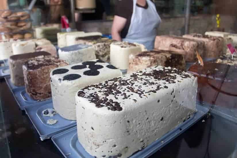 Various blocks of halva with different toppings displayed at a market stall, vendor in the background.