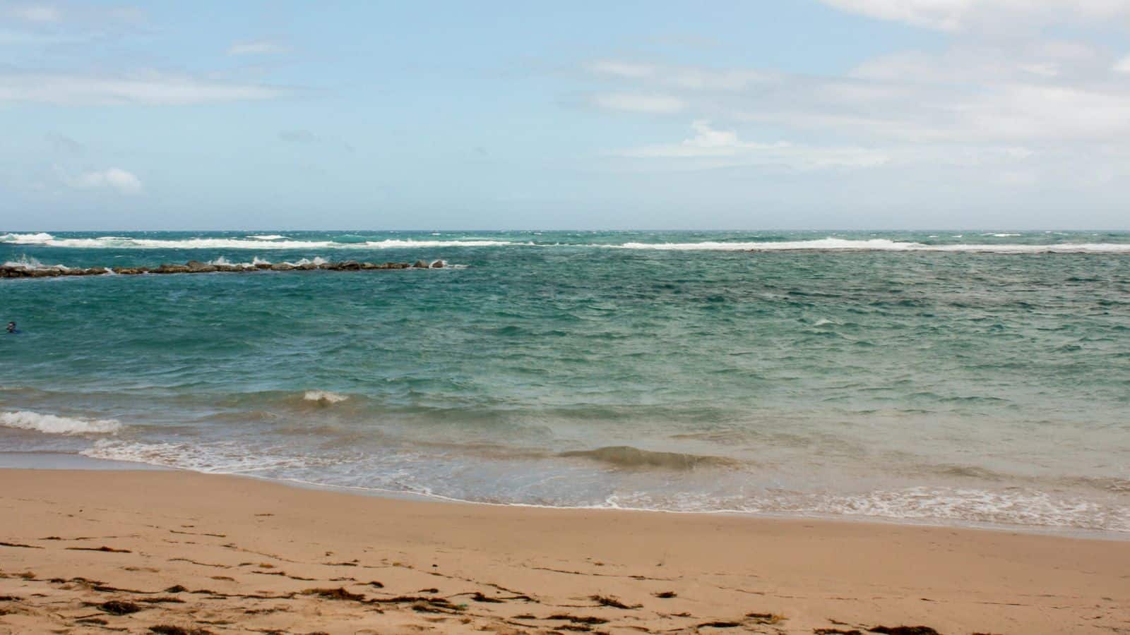 A sandy beach with scattered seaweed leads to blue-green ocean waves under a partly cloudy sky. A rocky breakwater extends into the water on the left side.