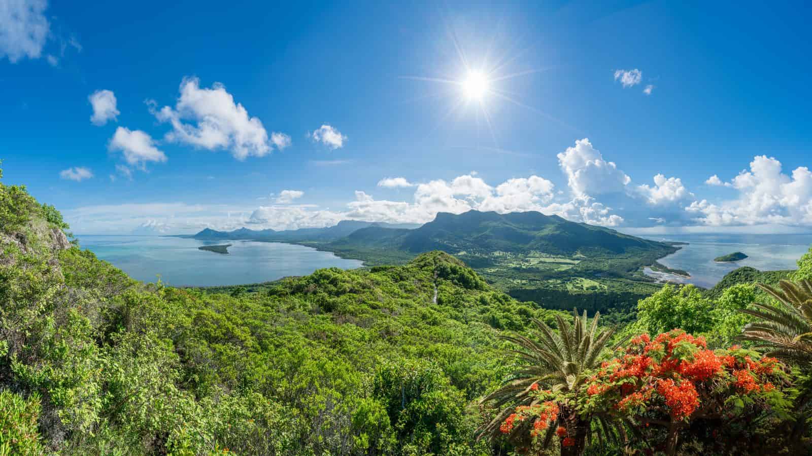 Lush green landscape with mountains, blue ocean, and bright sun in a clear sky.