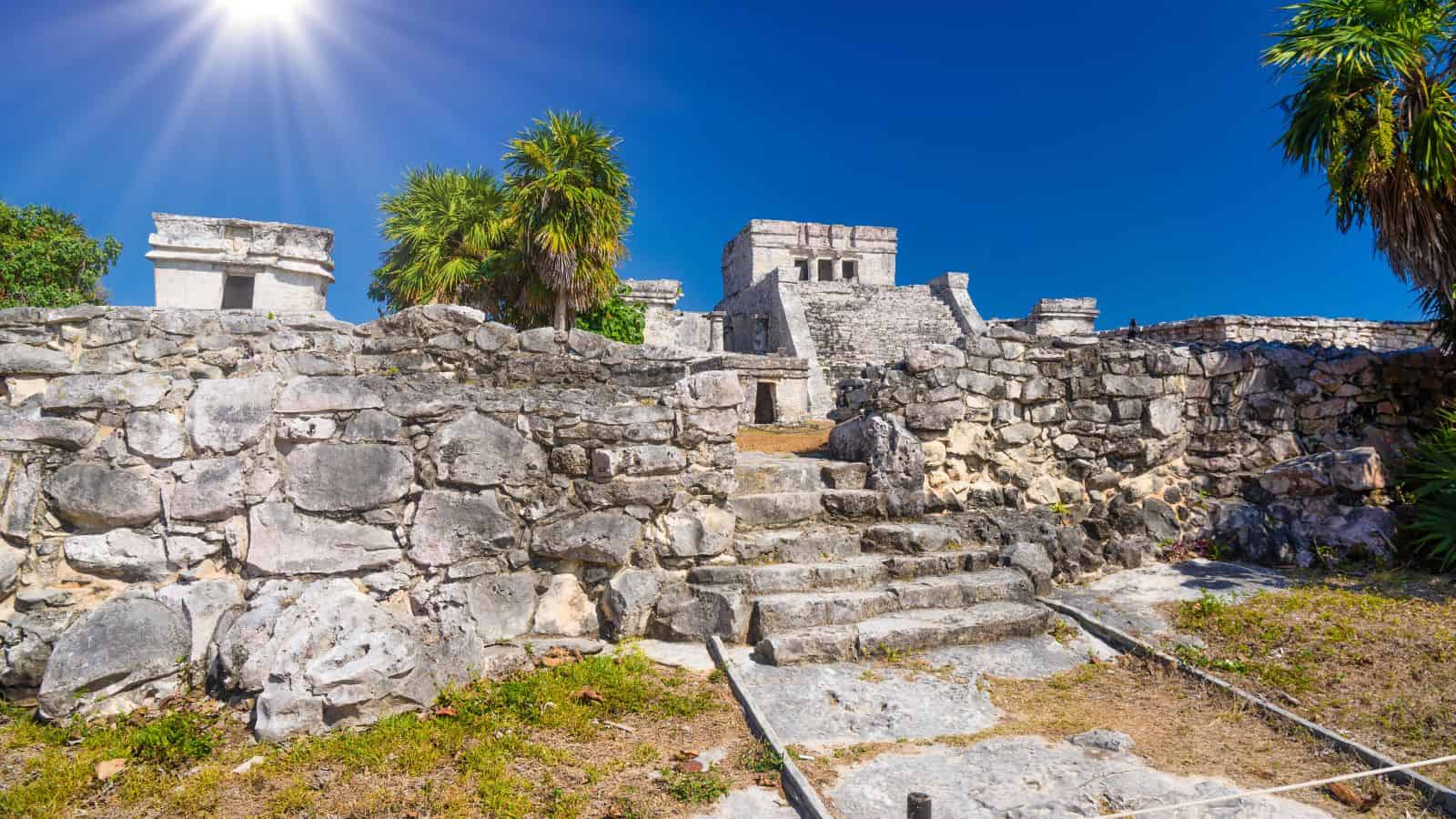 Ancient stone ruins of Tulum in Mexico under a bright sun and clear blue sky, with palm trees nearby.