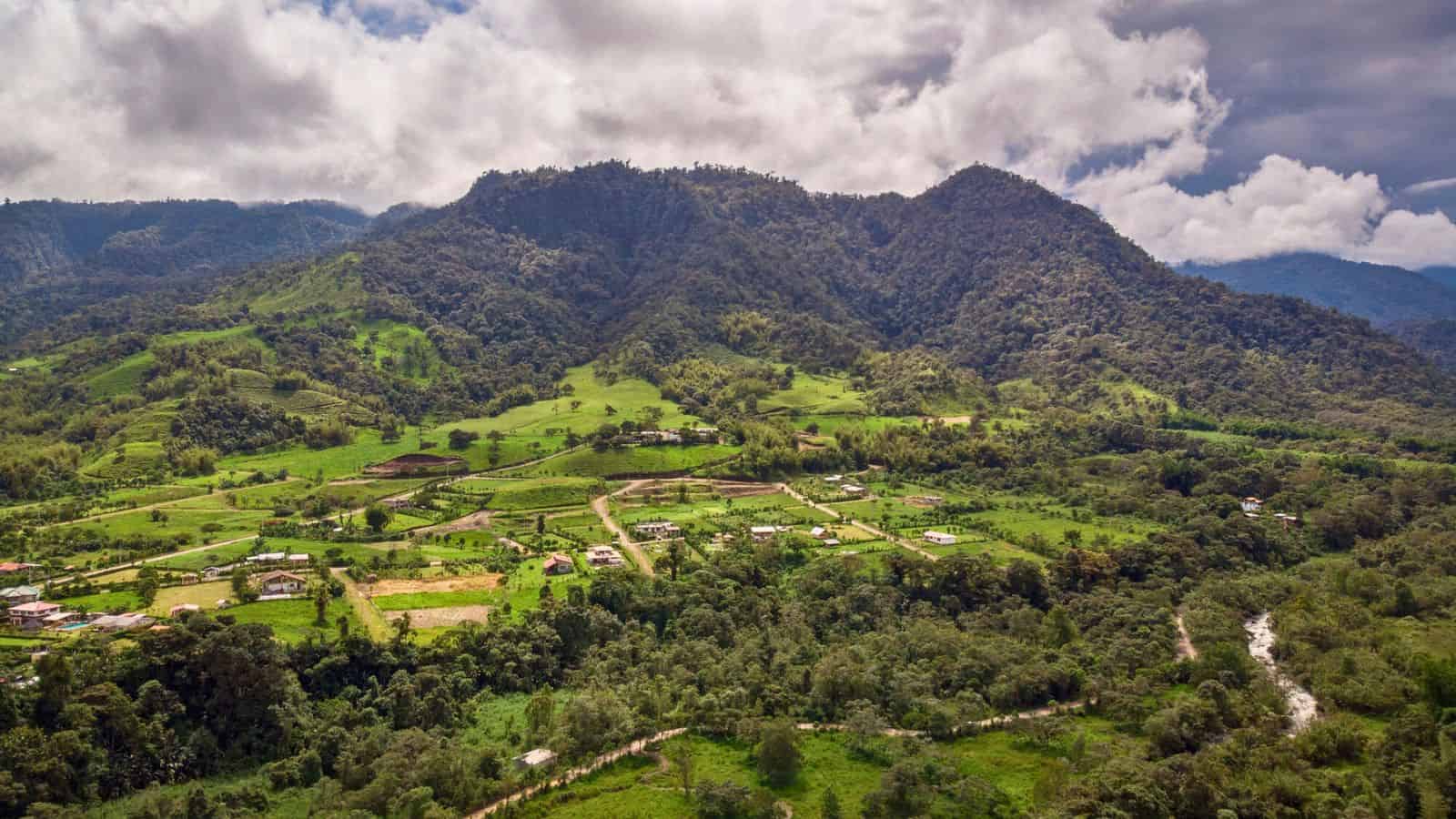 An aerial view of a green valley with scattered houses, trees, and mountains under a cloudy sky.