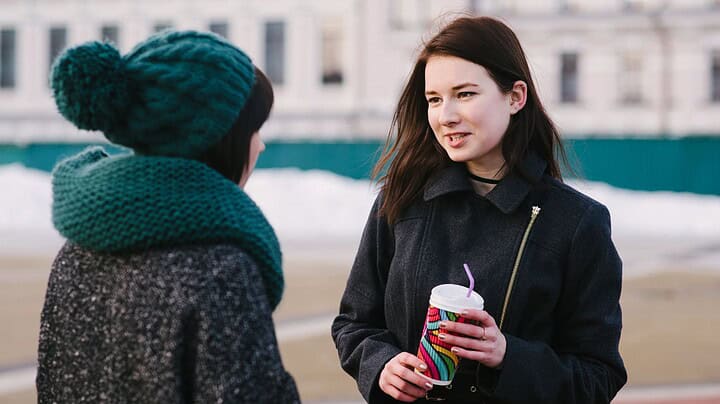 Two women talking outdoors in winter; one holds a colorful cup with a straw as they share Paris travel tips for their upcoming visit.