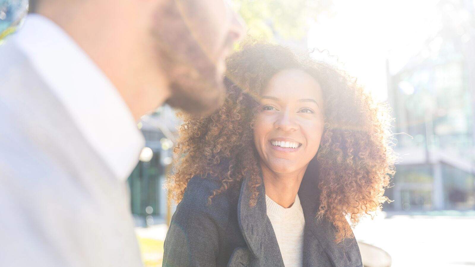A woman with curly hair sits outdoors, smiling at a man in the foreground. The image is bright with sunlight, and both people appear to be engaged in conversation. Trees and buildings are blurred in the background.