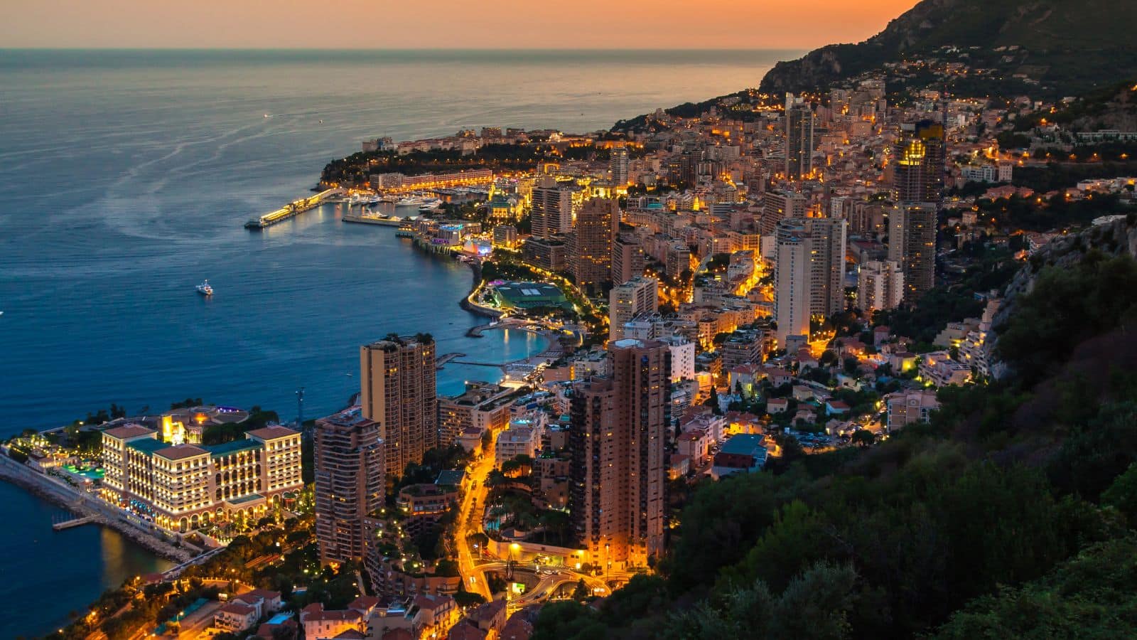 A coastal city at dusk with illuminated buildings, winding roads, and a harbor by the sea.