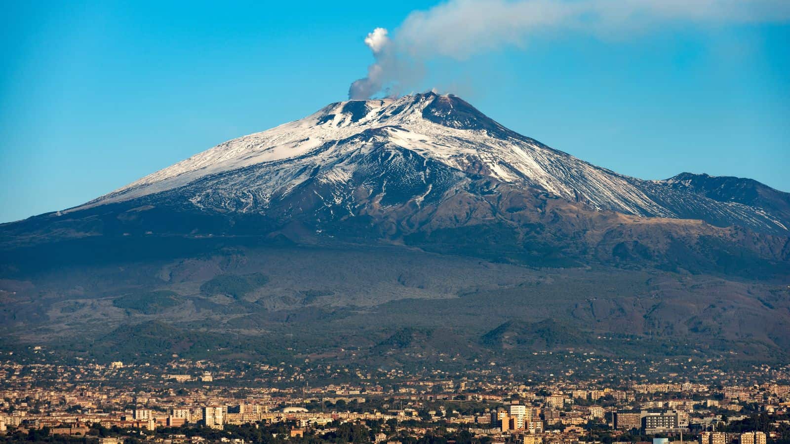 A snow-capped volcano emits a plume of smoke from its summit, with a densely built city and green vegetation spread out at the base under a clear blue sky.