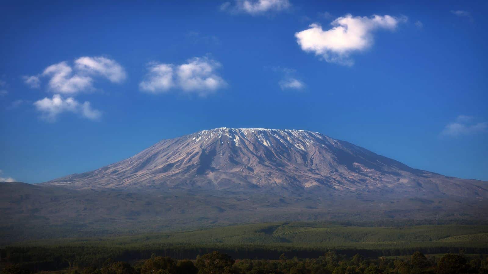 A mountain with a flat summit and patches of snow is seen under a blue sky with scattered clouds, with green forest at its base.