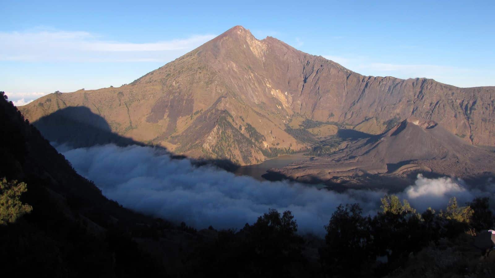 A mountain with a sharp peak rises above a valley filled with clouds, with rocky slopes and sparse vegetation visible under a clear blue sky. Foreground trees are silhouetted in shadow.