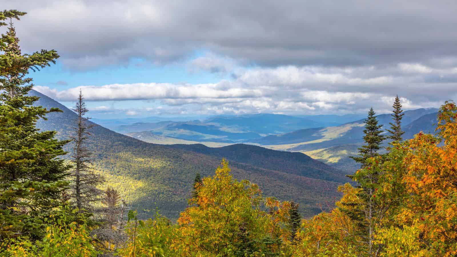 Mountain landscape with colorful autumn trees, rolling hills, and clouds in a bright blue sky.