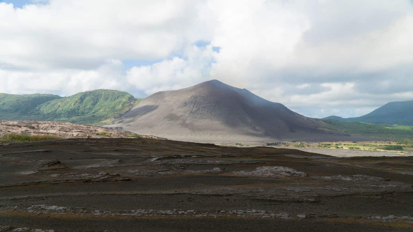A gray volcanic cone rises in the center of the image, surrounded by green hills and partially cloudy skies. The foreground shows dark, rocky terrain leading up to the base of the volcano.
