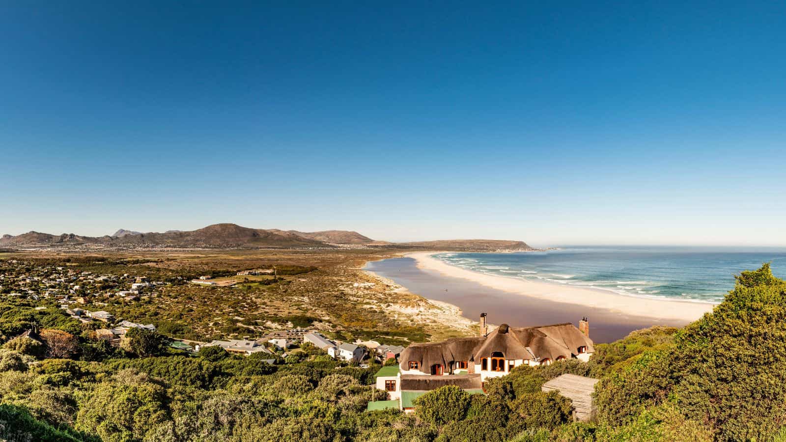 A wide beach stretches along the coastline with gentle waves, backed by green vegetation and scattered houses, under a clear blue sky with distant mountains in the background.