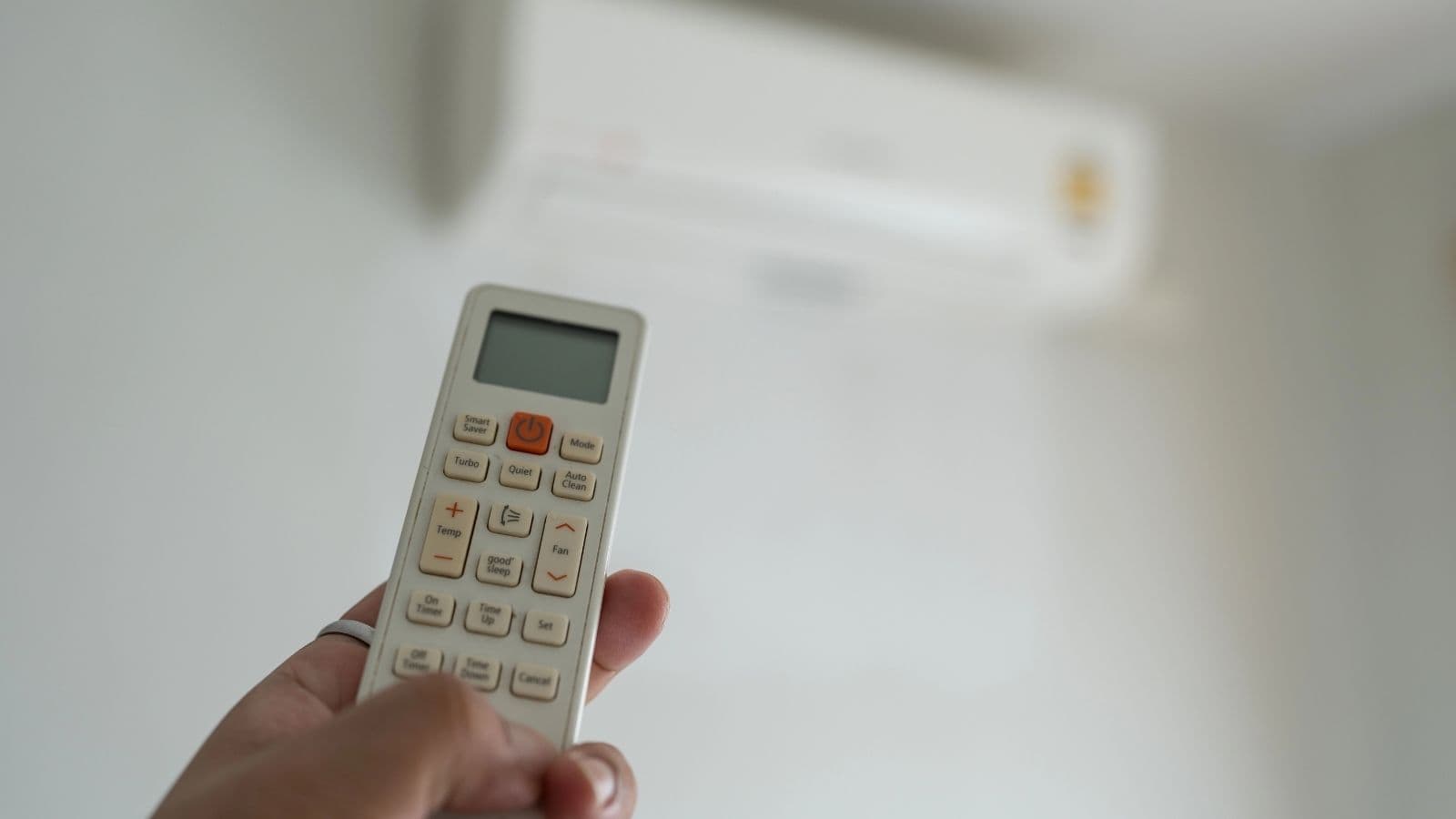A hand holding a remote control pointed at a wall-mounted air conditioner unit in a room with white walls—a handy travel tip for first-time travelers discovering European habits around climate control.