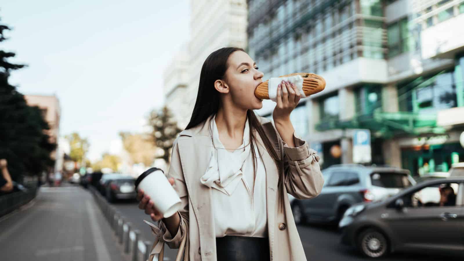 Woman in a city street eating a baguette and holding a coffee cup, with cars and buildings in the background.