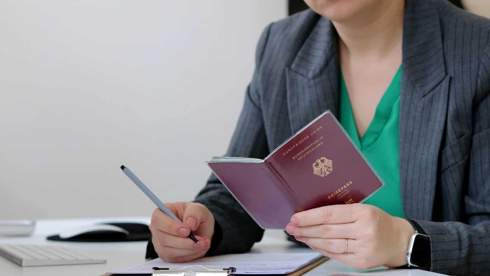 A person in a gray blazer and green blouse holds a burgundy passport and a pen, sitting at a desk with a clipboard, keyboard, and mouse. Their face is partially out of frame.