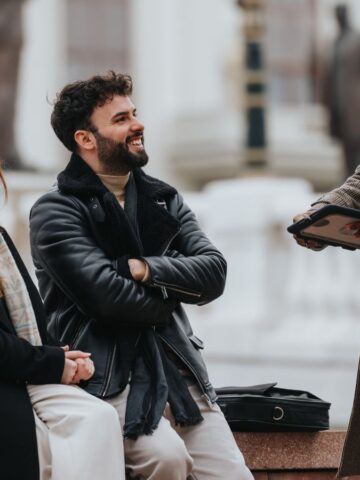 Three people talking outdoors in Paris; two locals seated and smiling, one standing and holding a tablet, all in winter clothes—perhaps sharing tips to avoid common tourist mistakes.