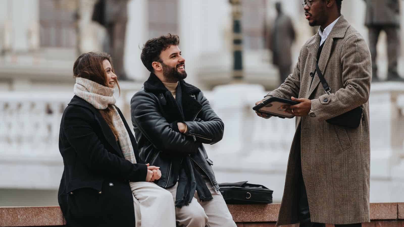 Three people talking outdoors in Paris; two locals seated and smiling, one standing and holding a tablet, all in winter clothes—perhaps sharing tips to avoid common tourist mistakes.