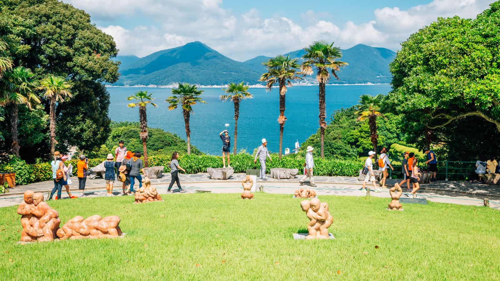 People walk among palm trees and abstract sculptures in a garden overlooking a blue lake and mountains.