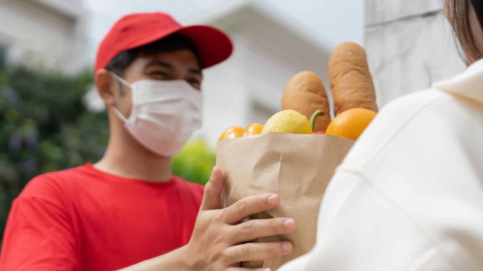 A masked delivery person hands a paper bag of groceries to someone outside.