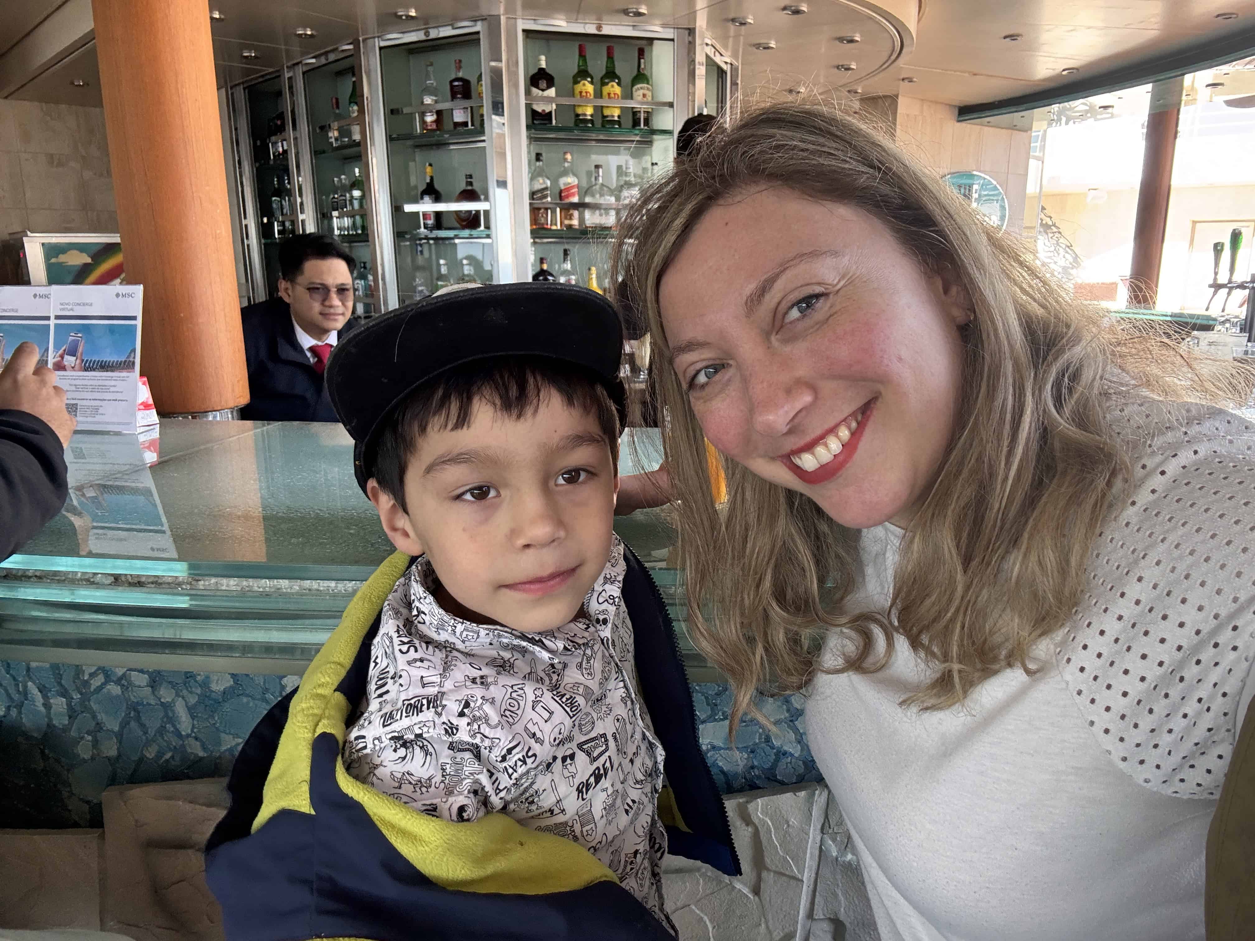 A smiling woman and young boy pose together at a bar with bottles and glasses in the background, capturing a joyful moment during their MSC Fantasia Dining experience.