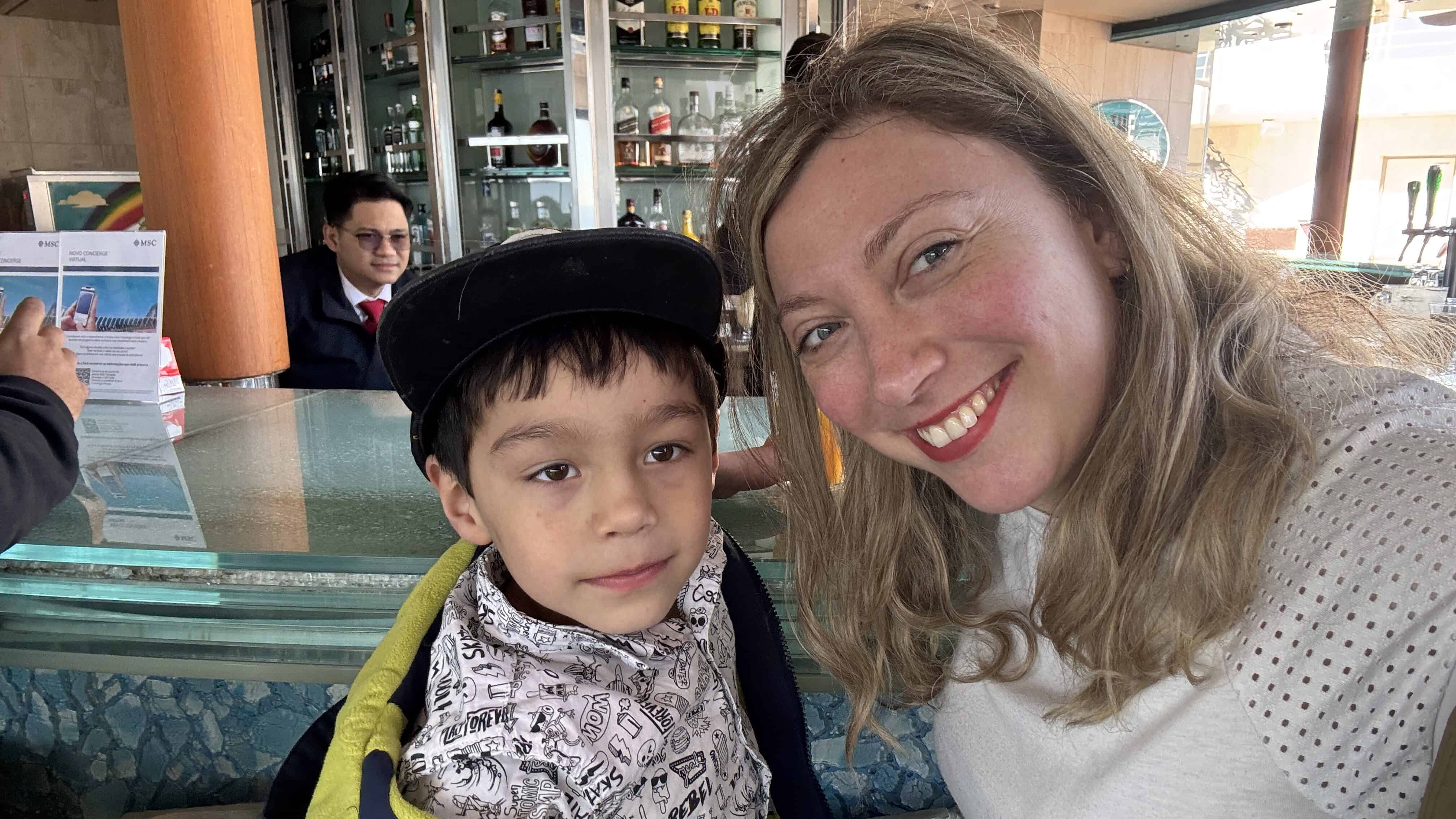 Woman smiling next to a young boy in a patterned shirt and cap at a bar with bottles in the background during their MSC Fantasia cruise—perfect for your 2025 Cruise Review.