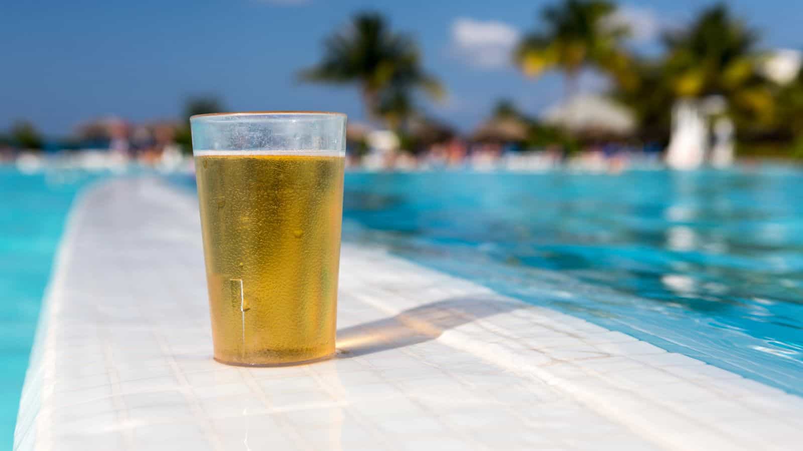 A clear plastic cup filled with light beer sits on the edge of a swimming pool&mdash;one of the many reasons travelers love relaxing at all-inclusive vacations, with palm trees and blurred people in the background on a sunny day.