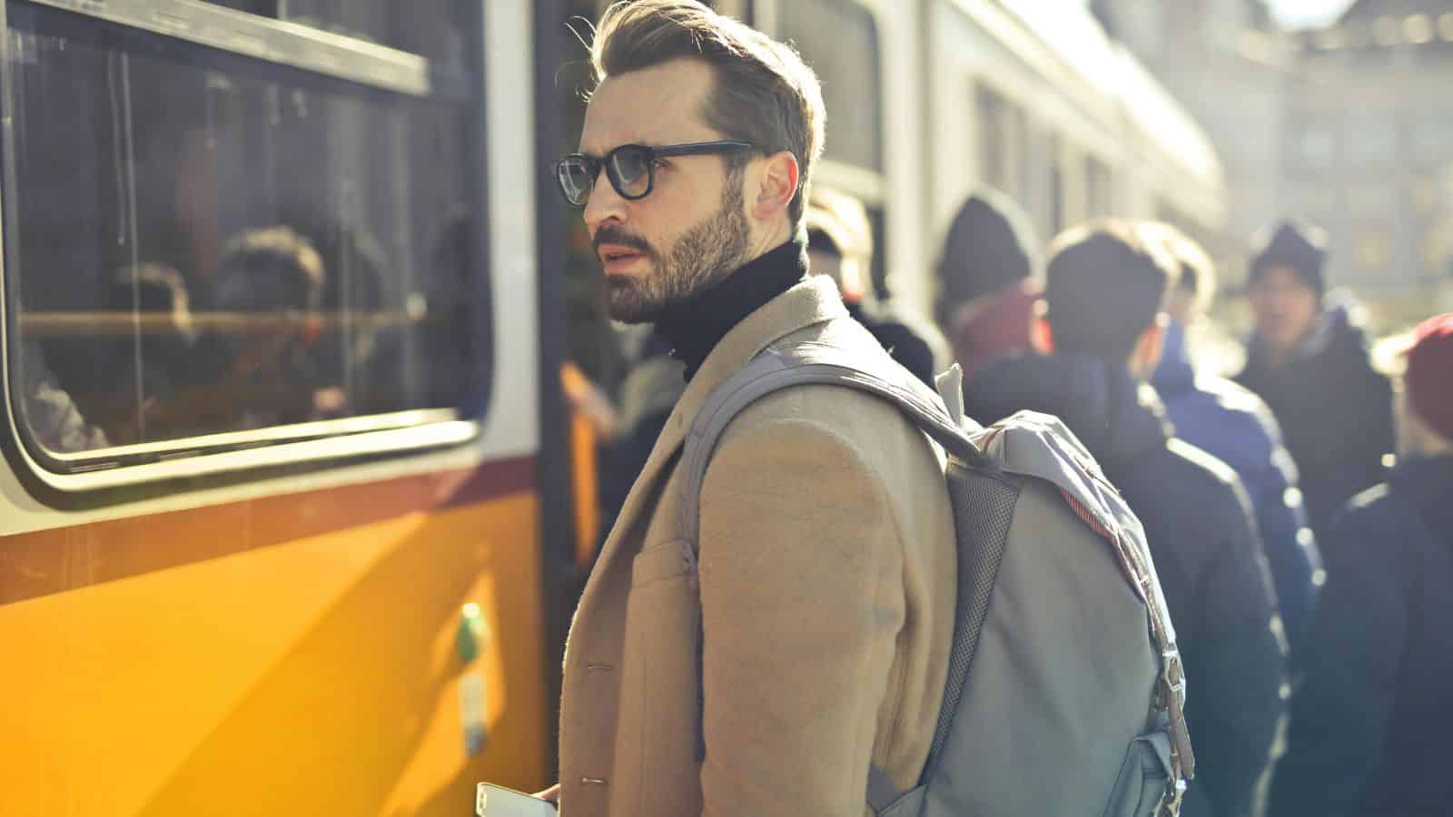 Man with glasses and backpack stands near a yellow bus, looking back, surrounded by other people.