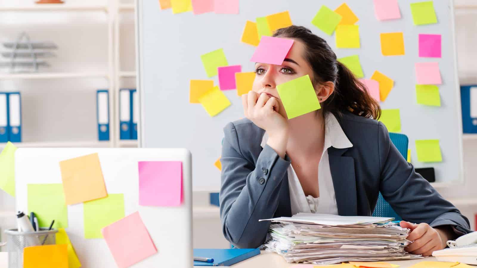 A woman at a cluttered desk, covered in sticky notes, looks overwhelmed and lost in thought.