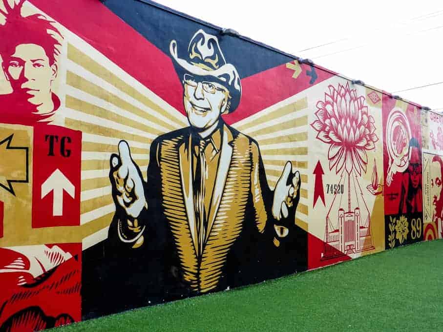 Colorful mural features a smiling man in a cowboy hat with hands open, surrounded by bold red and yellow graphics.