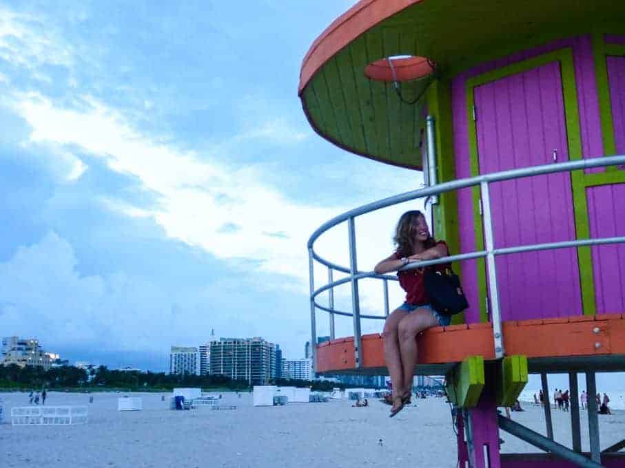 Woman sitting on a colorful lifeguard tower at the beach with city buildings in the background at dusk.